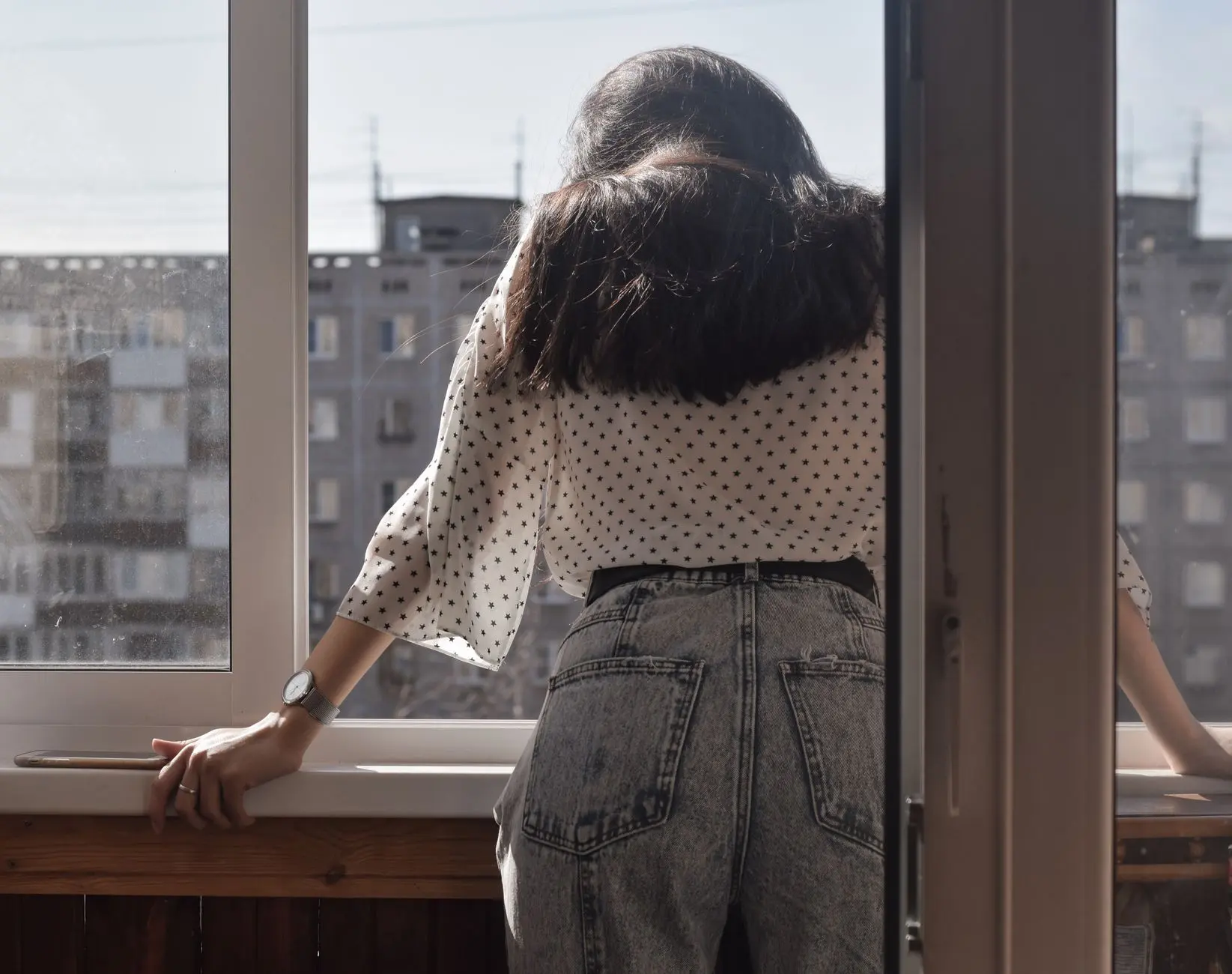 Una mujer a la moda en jeans contempla la ciudad desde una ventana, capturando la vida urbana moderna.