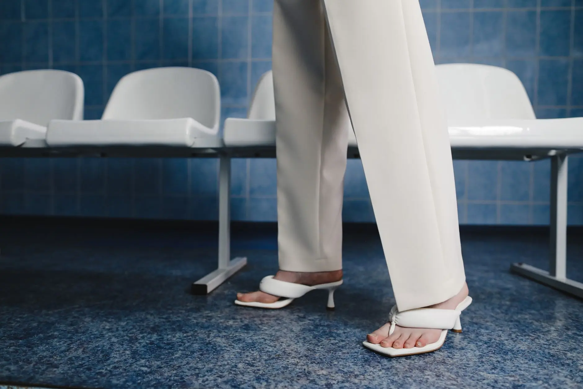 Imagen de moda minimalista de una mujer con pantalones blancos y sandalias de pie en el interior junto a asientos.