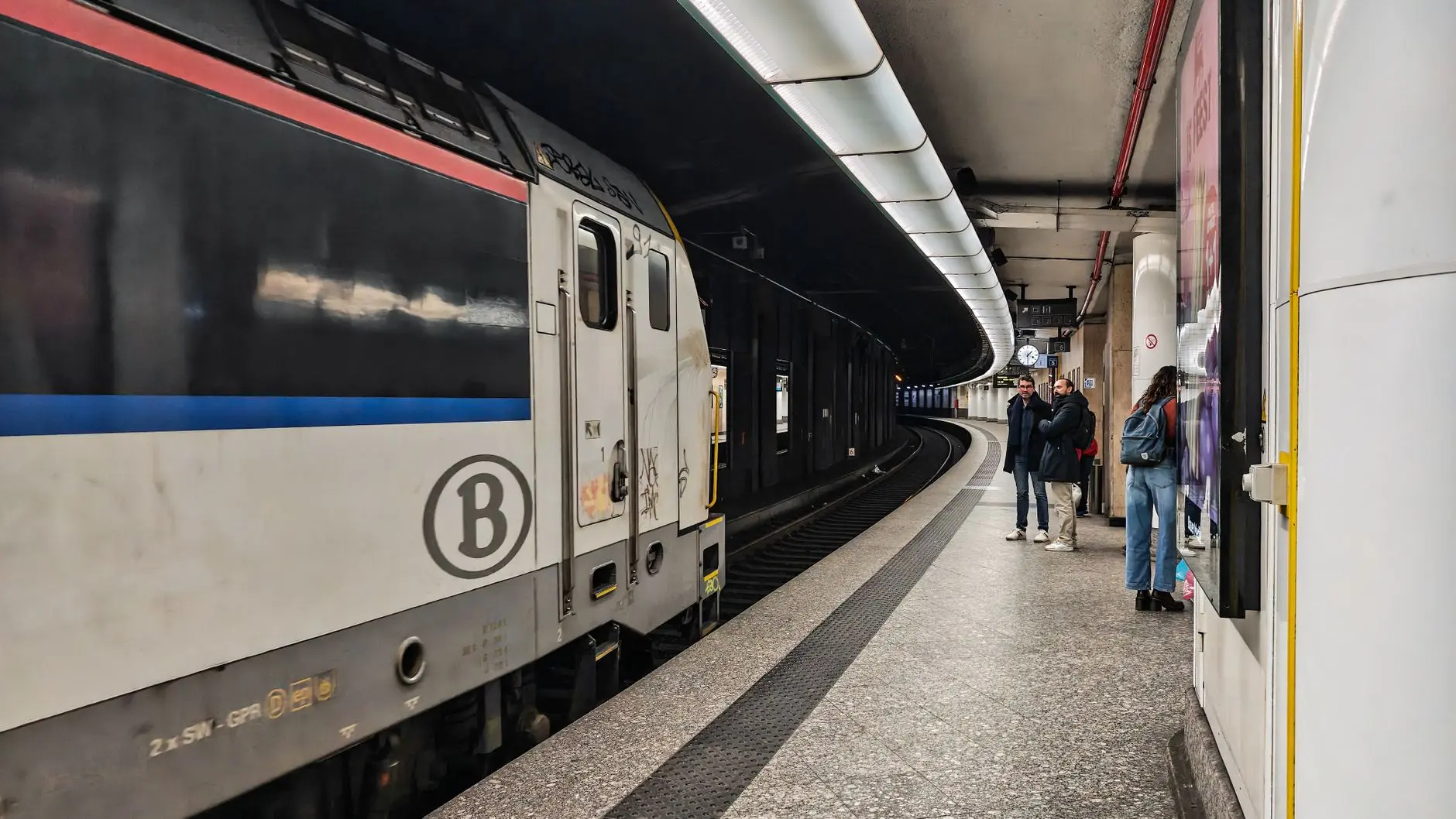 Personas esperan en una concurrida estación de tren en Bruselas, con un tren acercándose a la plataforma.