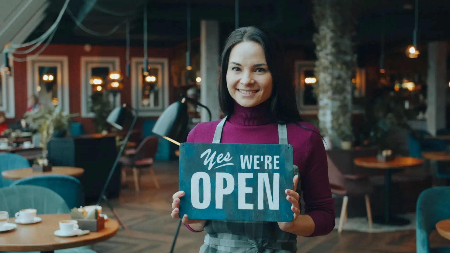 El dueño de una cafetería, con una sonrisa radiante, sostiene un cartel de "Abierto" en el interior de un local acogedor y con una iluminación cálida.